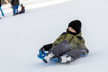 Bir çocuk kızağa biniyor. Kışlık ceketli bir çocuk tepeden aşağı iniyor.. 
