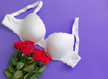 Top view white lace lingerie with red roses on violet background. Set of woman essential underwear or accessory. Flat lay, beauty blog or social media minimal concept. Present for Valentines day