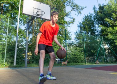 Cute young teenager in red t shirt with a ball plays basketball on court. Teenager dribbling the ball,  running in the stadium. Sports, hobby, active lifestyle for boys