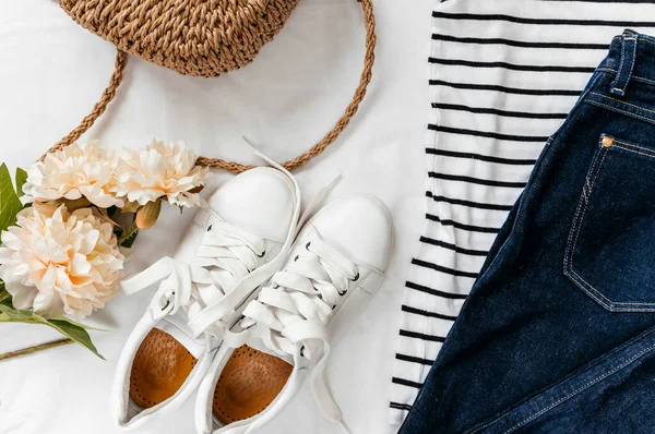 Blue jeans, t shirt, bag and white sneakers on white bed. Trendy clothes. Women\'s casual spring summer outfit. Fashion concept. Flat lay, top view