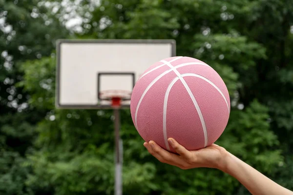 Pink basketball ball in man\'s hands in front of a basket outdoors. Minimal sport background. Sports gear, close up