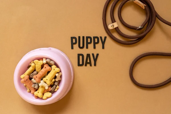 Close up of a dog bowl with bones, leather leash on brown background. Title Puppy day in the middle. Dog accessories, rubber toys. Minimalistic background.