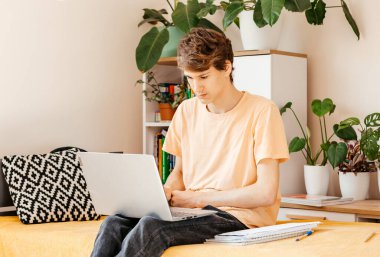 Evdeki kanepede dizüstü bilgisayarla çalışan konsantre bir genç. Gülümseyen Çocuk bilgisayarla ödev yapıyor. Çocuklar için uzaklık ve çevrimiçi eğitim