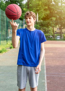 Tişörtlü şirin çocuk şehir parkında basketbol oynuyor. Aktif gençler kırmızı topla dışarıda oyun oynuyorlar. Hobi, aktif yaşam tarzı, çocuklar için spor.