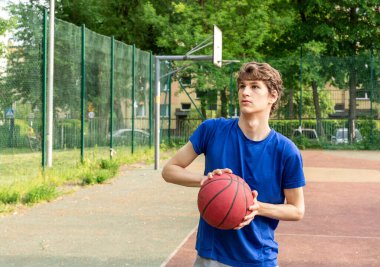 Tişörtlü şirin çocuk şehir parkında basketbol oynuyor. Aktif gençler kırmızı topla dışarıda oyun oynuyorlar. Hobi, aktif yaşam tarzı, çocuklar için spor.
