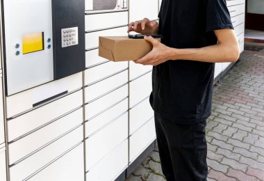 Posta terminalinin yanında elinde kutu olan bir adam. Paket teslimat makinesi. Elinde bir karton kutu tutan kişi. Posta ve posta servisi, online alışveriş, e-ticaret konsepti