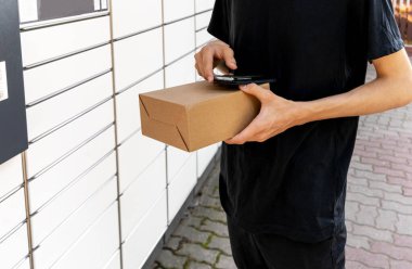 Posta terminalinin yanında elinde kutu olan bir adam. Paket teslimat makinesi. Elinde bir karton kutu tutan kişi. Posta ve posta servisi, online alışveriş, e-ticaret konsepti