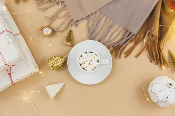 Cup of coffee, gift box and knitted sweaters, led lights on beige background. Hygge style, winter concept. Cozy home desk. New year and Christmas celebration mockup. Christmas still life