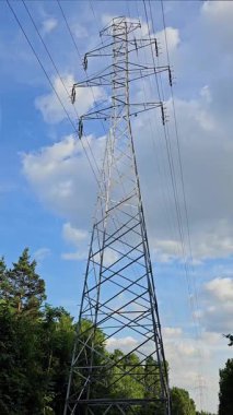 Uzun bir çelik güç kulesi, kabarık bulutlarla kaplı parlak mavi gökyüzüne doğru yükseliyor. Yeşilliğin etrafını sarmak manzaraya doğal bir dokunuş katar..