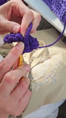 Bir insan rahat ve güneşli bir öğleden sonra otururken bir kanca ve iplik kullanarak ustalıkla mor bir çiçek örer. Parlak renk doğal çevreye karşı göze çarpar..