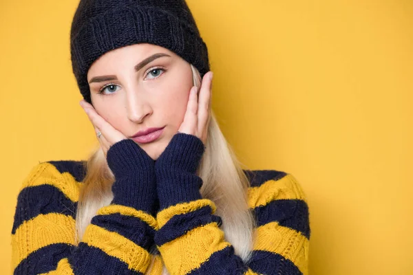 Head shot of young beautiful woman with hands keeping on cheek, model wearing woolen cap and sweater, isolated on yellow background. Close up face of cute girl. Studio shot