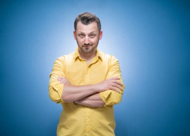 Mavi arka planda çapraz kolları olan, sarı gömlekli, ciddi yakışıklı bir adam. Sakallı adam kameraya bakıyor. Stüdyo çekimi