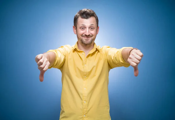 親指を示す笑顔の若い男は 青の背景に概念を嫌い 黄色のシャツのドレス スタジオショット — ストック写真