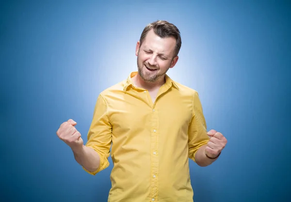 青い背景の上に拳を握りしめ 黄色のシャツに身を包んだ興奮した若い男 はいコンセプト 良い知らせだ 楽しい男は成功を祝う スタジオショット — ストック写真