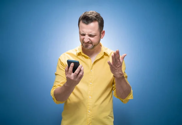 Nervous man holding mobile phone over blue background, dresses in yellow shirt. No Wi-fi concept. Bad connection, broken network low signal. Studio shot