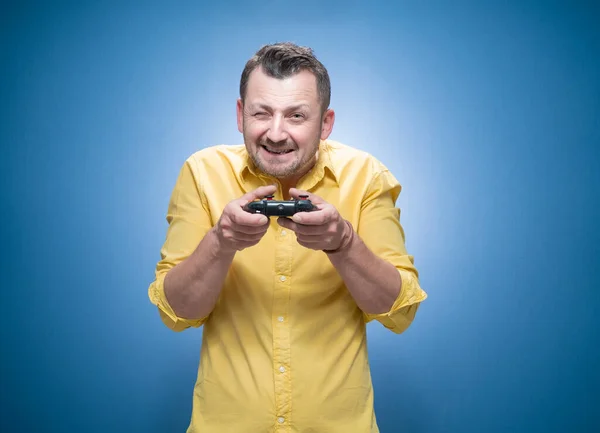 Cheerful man with eyes closed holding video games joystick over blue background, dresses in yellow shirt. Guy have fun and holding game controller. Studio shot