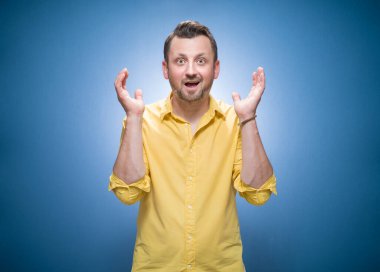 Amused young man with opened hands over blue background, dresses in yellow shirt. Yes concept. Good news. Amusing guy celebrates success. Studio shot