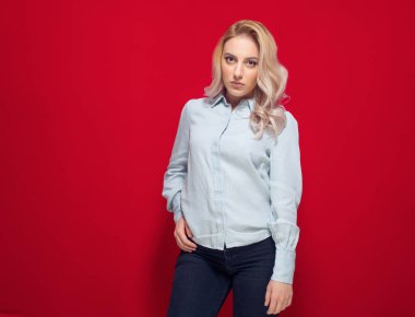 Beautiful woman with hands in pockets looking in camera while standing, isolated on red background. Attractive girl. Studio shot