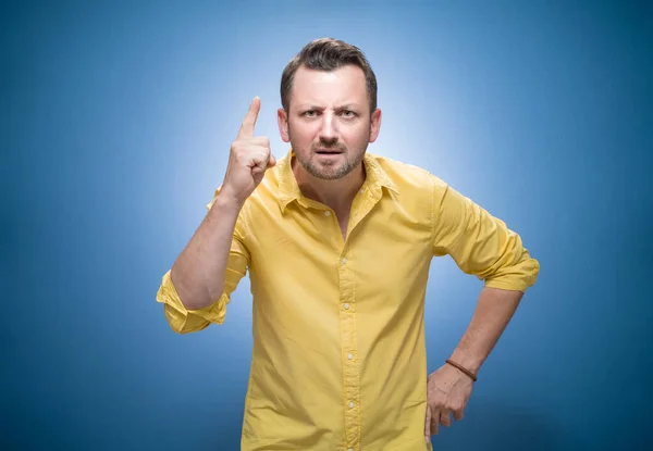 Angry man pointing finger up with the index finger a showing problem over blue background, dresses in yellow shirt. Fury guy screaming. Studio shot