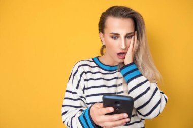 Cep telefonu olan korkmuş genç bir kadın, sarı arka planda izole edilmiş. Şok olmuş kız elinde akıllı telefon tutuyor. - İnanılmaz. Stüdyo çekimi