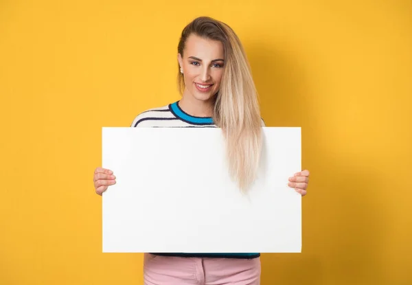 Promoting women holding white board, isolated on yellow background. Amused girl showing blank empty paper billboard with blank space for text. Studio shot