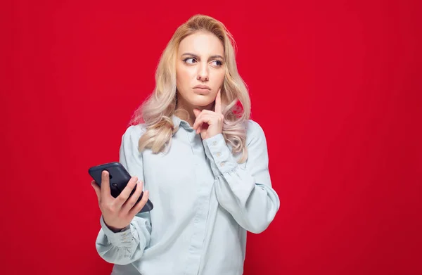 试想一下 一个时髦的女孩在手机上浏览 在红色背景下被隔离 一个拿着手机思考的年轻女人工作室拍摄 图库图片