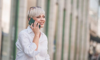 40 'lı yaşlarda sarışın bir kadın cep telefonuyla şehirdeki bankta konuşuyor ve dinleniyor. Yetişkin kadın akıllı telefondan konuşuyor. Dışarıda, dışarıda.