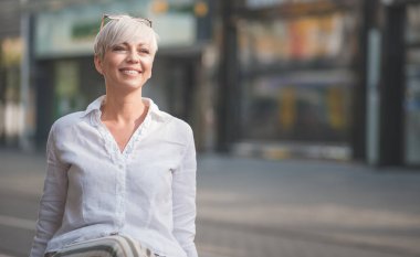 Sarışın, kısa saçlı, güzel bir kadın. Bankta otururken beyaz yaz gömleği giymiş neşeli bir kadın. Açık hava, bulanık doğa geçmişi.