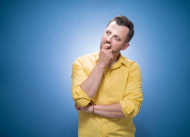 Genç adam yukarı bakıyor ve düşünüyor. Yüzünde gülümseme ifadesi olan son derece ilham verici bir adamın portresi, mavi arkaplanda metin için kopyalama alanı, sarı gömlekli elbise. Stüdyo çekimi