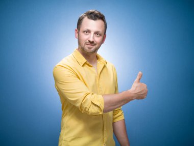 Baş parmakları havada bir adam, mavi arka plan üzerinde konsept gibi, sarı gömlekli. Evet, el kol hareketi, iyi iş çıkarmanın şarkıları. Yarı uzunlukta memnun bir adam. Stüdyo çekimi