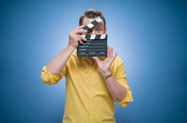 Genç adam mavi arka planda el çırpma tahtası tutuyor, sarı gömlekli. Stüdyo çekimi