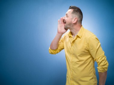 Mavi arka planda bağıran, sarı gömlekli bir adam. Sakallı adamın yandan görünüşü bir şey söylüyor. Boşluğu kopyala - metin için boşluk. Stüdyo çekimi