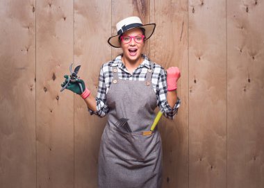 Heyecanlı bahçıvan kadın elinde budama makası, ağzı açık bağırıyor ve stüdyoda tahta arka planda yumruklarını sıkıyor. Şaşırmış kadın çiftçi başarıyı kutluyor. Stüdyo çekimi