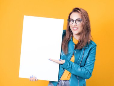 Mavi deri ceket giymiş, sarı arka planda soyutlanmış, beyaz tahta tutan gülümseyen genç kadınlar. Boş ilan panosu ve mesaj atmak için boş yeri olan boş bir kız. Stüdyo çekimi