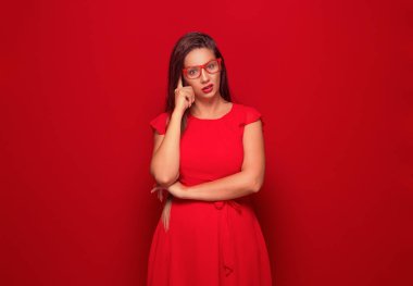 Düşünceli genç bir kadın, kırmızı arka planda izole edilmiş. Şakağa parmağını dayayıp kaşlarını çatan yalnız kız çapraz kollarla karışık. Stüdyo çekimi