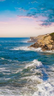 Kıyı ve deniz, güzel kayalık kıyılar. Taşlaşmış sahil üzerinde gün batımı ve kayalık kıyılara vuran tatlı okyanus suyu. Huzursuz mavi deniz. Atlantik Okyanusu - Portekiz