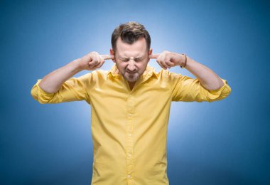 Gürültü - yüksek. Genç, sinirli, stresli bir adamın belden yukarı portresi kulaklarını kapatan, mavi arka planda gözlerini kapatan, sarı gömlekli bir adam. Stüdyo çekimi