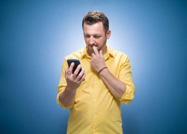 Mavi arka planda akıllı telefonu olan, sarı tişörtlü, uzun boylu, endişeli bir genç. Haberler kötü. Stüdyo çekimi