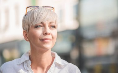 Sarışın, kısa saçlı, güneş gözlüğü takan orta yaşlı güzel bir kadın. Beyaz yaz gömleği giymiş kadın profili. Açık havada, şehrin bulanık sokaklarında..