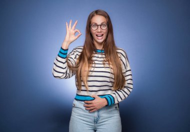 Şaşırmış genç bir kadın, mavi arka planda izole edilmiş parmağıyla mükemmel bir işaret gösteriyor. Evet ya da mükemmel jestler, vücut dili konsepti. Stüdyo çekimi