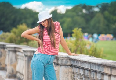 Genç ve güzel bir kadın ayakta ve aşağı bakıyor, dışarıda - bulanık arka plan - bokeh. Güzel bir kızın beli beyaz şapka ve yaz kıyafetleri giyer. Gün ışığı