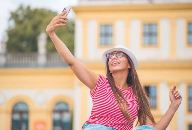 Moda kızı elinde akıllı telefon tutuyor ve dışarıda, arka planda bulanık bir şekilde selfie çekiyor. Genç bir kadın akıllı telefon kullanıyor ve kendi fotoğrafını çekiyor. Gün ışığı
