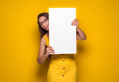 Sarı arka planda izole edilmiş beyaz tahta tutan gülümseyen genç kadınlar. Boş ilan panosu ve mesaj atmak için boş yeri olan güzel bir kız. Stüdyo çekimi