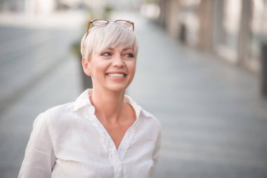 Sarışın, kısa saçlı, güzel bir kadın. Beyaz yaz tişörtü giymiş neşeli bir kadın. Açık havada, şehrin bulanık sokaklarında..