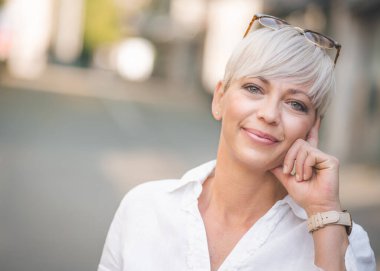 Avrupalı mutlu sarışın yetişkin kadının yakın plan portresi. Beyaz yaz gömleği giymiş güzel bir kadın yüzü. Açık havada, şehrin bulanık sokaklarında..