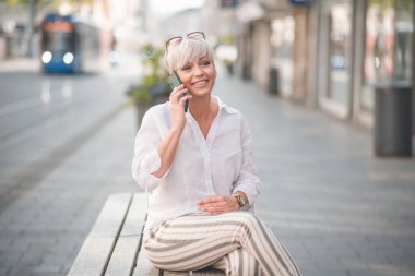 Cep telefonuyla konuşan, kentteki bankta dinlenen orta yaşlı güzel bir kadın. Yetişkin kadın akıllı telefondan konuşuyor. Dışarıda, dışarıda.