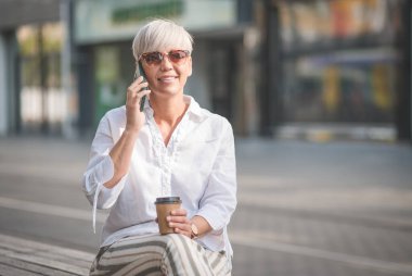 Cep telefonunda güneş gözlüklü şık sarışın kadın kentteki bankta konuşuyor ve dinleniyor. Yetişkin kadın akıllı telefondan konuşuyor. Dışarıda, dışarıda.