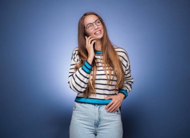 Akıllı telefondaki neşeli kız konuşuyor, mavi arka planda izole edilmiş. Keyif ve cep telefonu. Stüdyo çekimi