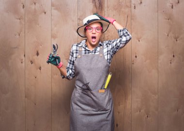 Panik halinde, elinde budama makası tutan genç bahçıvan kadın stüdyoda ahşap arka planda sana bakıyor. Yanlışlık. Haberler kötü. Stüdyo çekimi