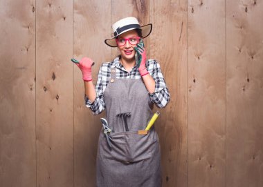 Memnun genç bahçıvan kadın stüdyodaki ahşap arka planda akıllı telefondan konuşuyor. Akıllı telefonu kullanan mutlu bir kadın çiftçi. Stüdyo çekimi